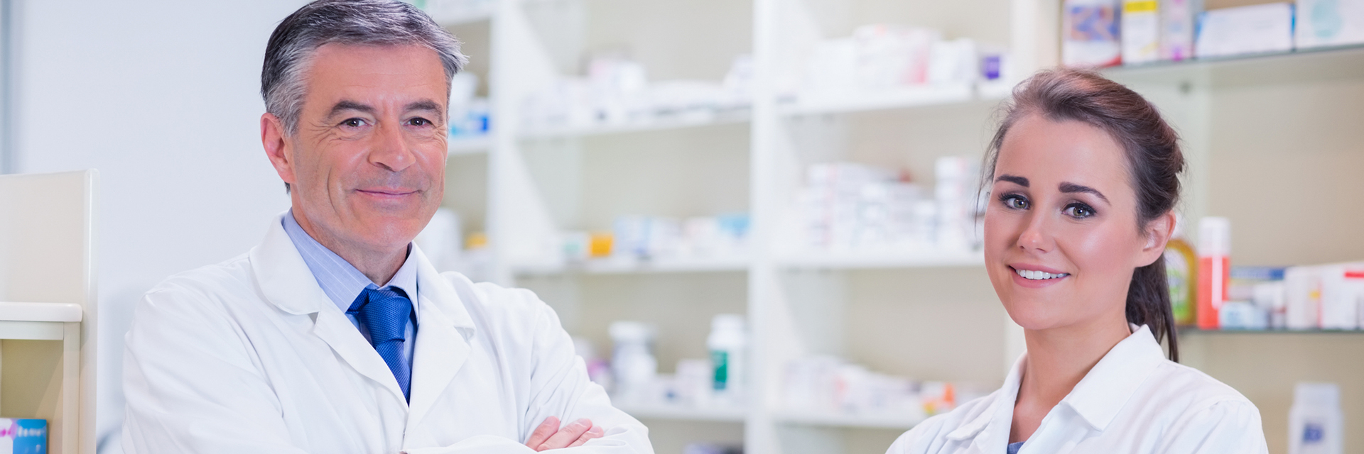 Male and female pharmacist both smiling at the camera with pharmacy shelves in background